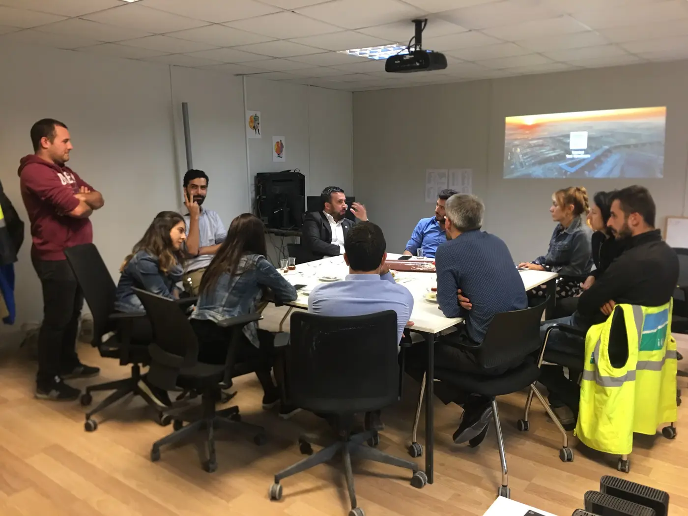 Toplantı masasında ekip ve projeksiyonda gösterilen şantiye görüntüsü
